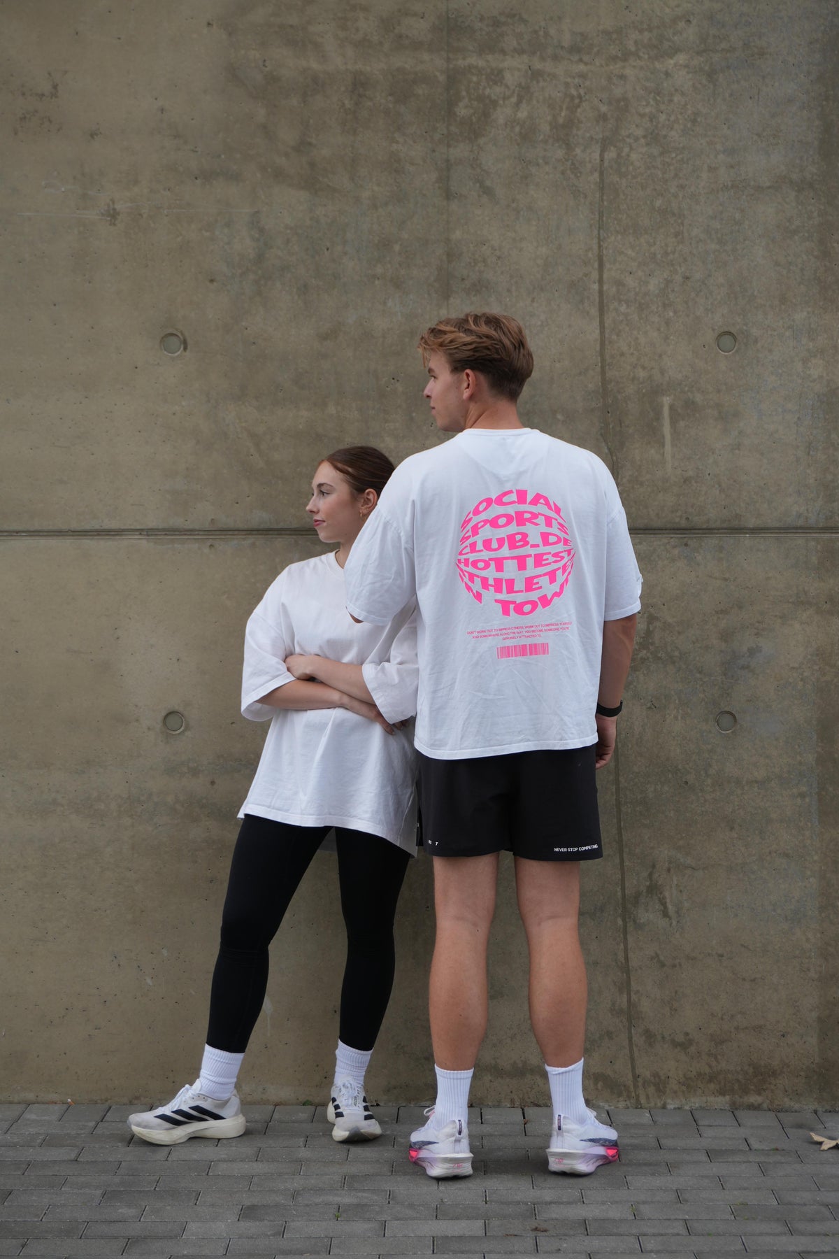 HOT-TEE 1.0 zu zweit vor Betonwand: Front clean und pinker Backprint – Social Sports Club
